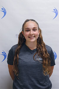 Corinne H. portant un t-shirt gris devant un fond blanc avec logo.