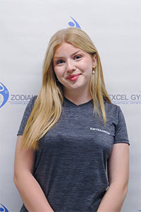 Portrait de Jasmine B., portant un t-shirt gris devant un logo de gymnase.